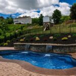 freeform shaped pool with a waterfall feature and patio furniture at the base of a sloped backyard