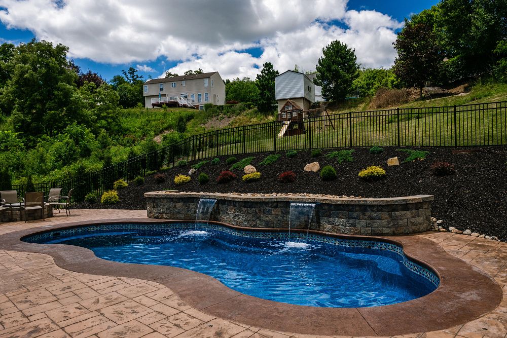 freeform shaped pool with a waterfall feature and patio furniture at the base of a sloped backyard