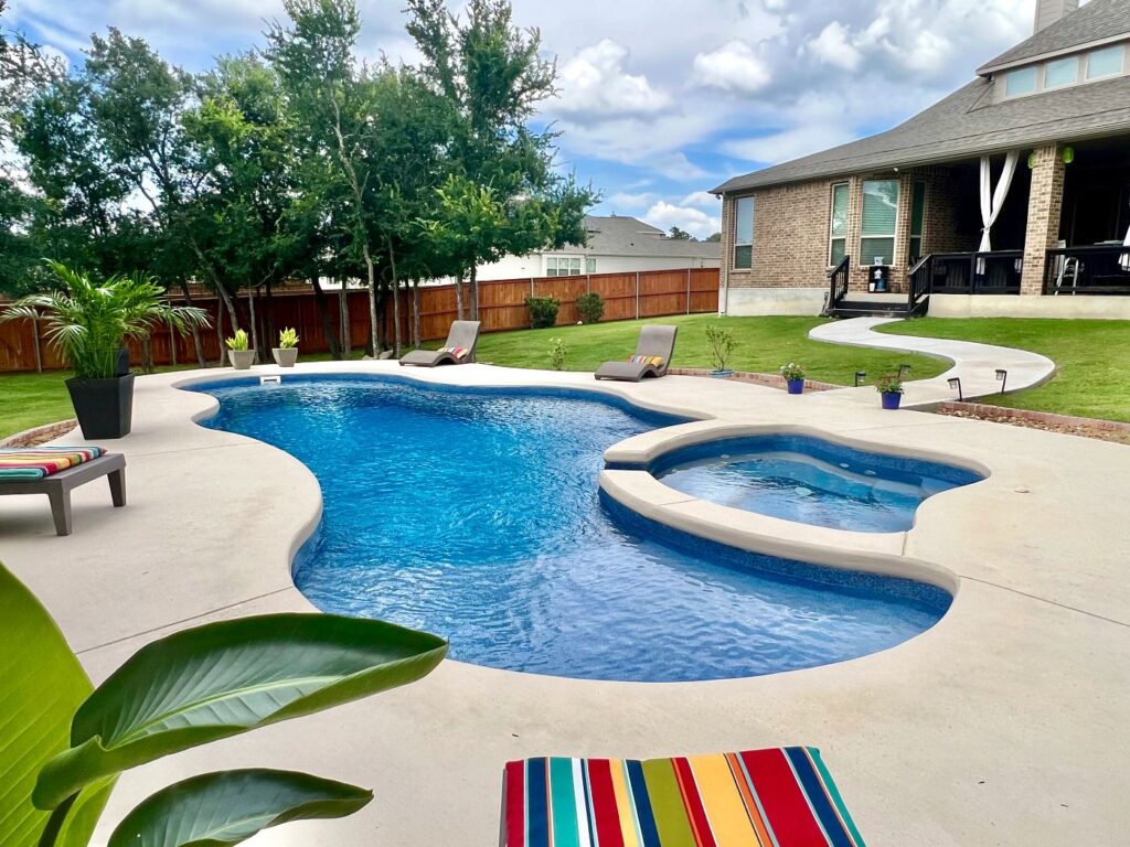 Freeform fiberglass pool and integrated elevated spa with light concrete decking in a sunny Texas backyard.