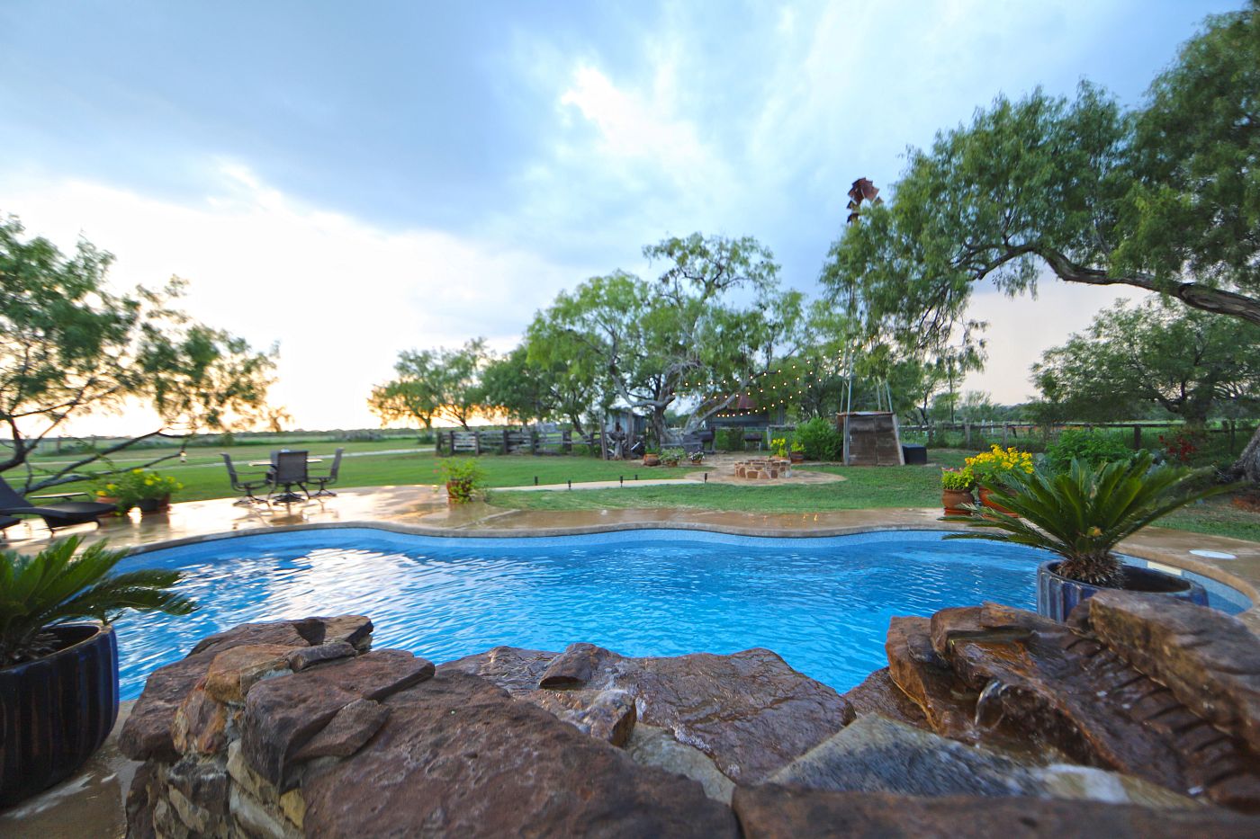 Large backyard fiberglass pool framed by rocks, greenery, and a spacious lawn.