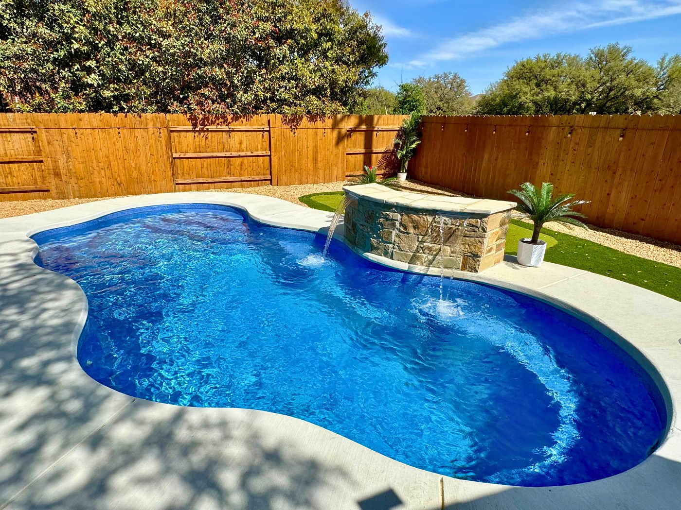 Freeform fiberglass pool with raised water fountains, surrounded by wood fencing and greenery.