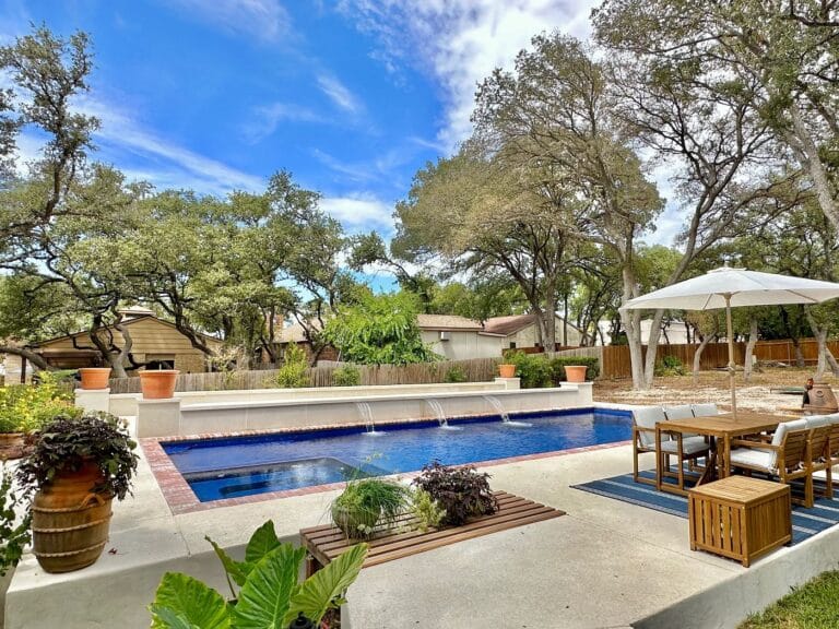 Backyard fiberglass pool with stone patio, surrounded by trees and outdoor seating.