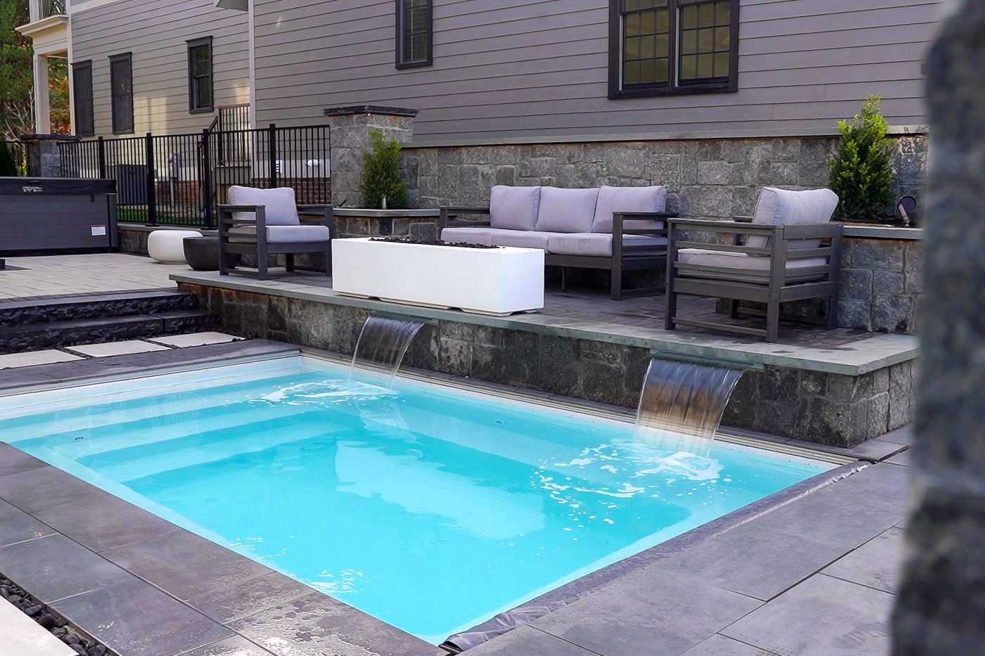 Compact rectangular plunge pool with built-in waterfall, framed by a stone patio and modern seating.