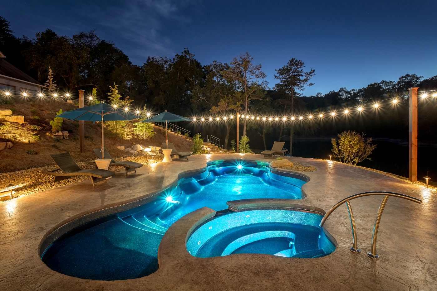 Freeform fiberglass pool and elevated integrated spa lit up at night by warm string lights and blue underwater pool lighting.
