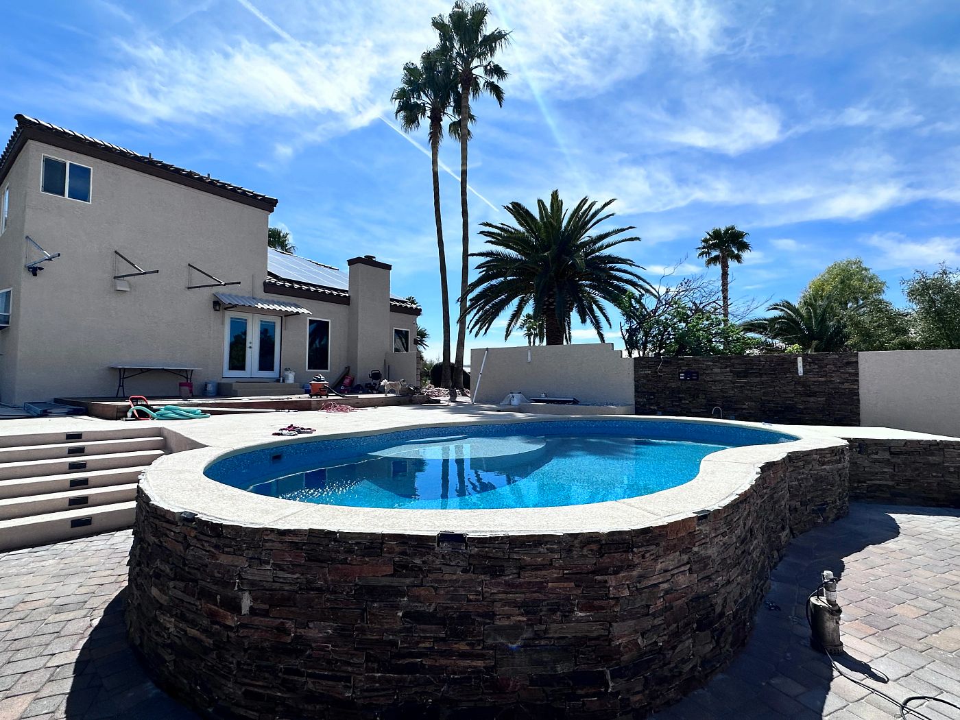 Freeform semi-above ground vinyl liner pool with stone masonry retaining wall and palm trees in a sunny Florida backyard.